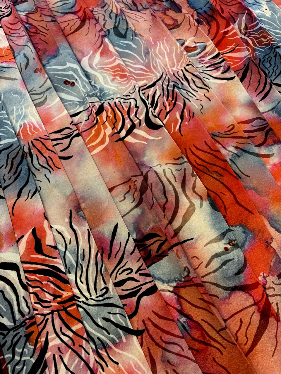 A close-up of a fabric displaying a vibrant pattern, featuring abstract floral and linear designs in shades of red, orange, grey, and black. The fabric appears draped or layered, enhancing the texture and depth of the pattern.