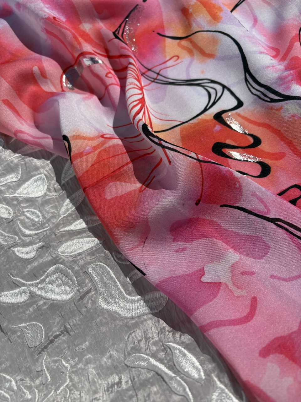 A close-up of a patterned fabric featuring vibrant pink and orange abstract designs, layered over a textured silver surface. The fabric's sheer quality allows the colours and patterns to blend subtly with the underlying texture.