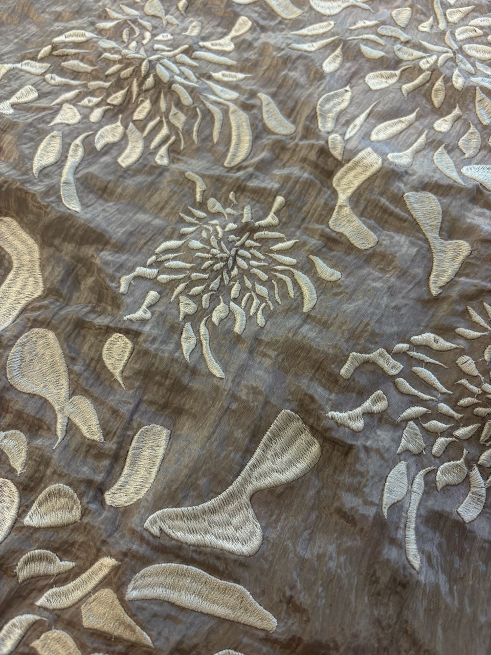 A textured fabric featuring an intricate floral embroidery design in cream on a brown background. The flowers are stylised, with flowing shapes and curves creating an elegant pattern.