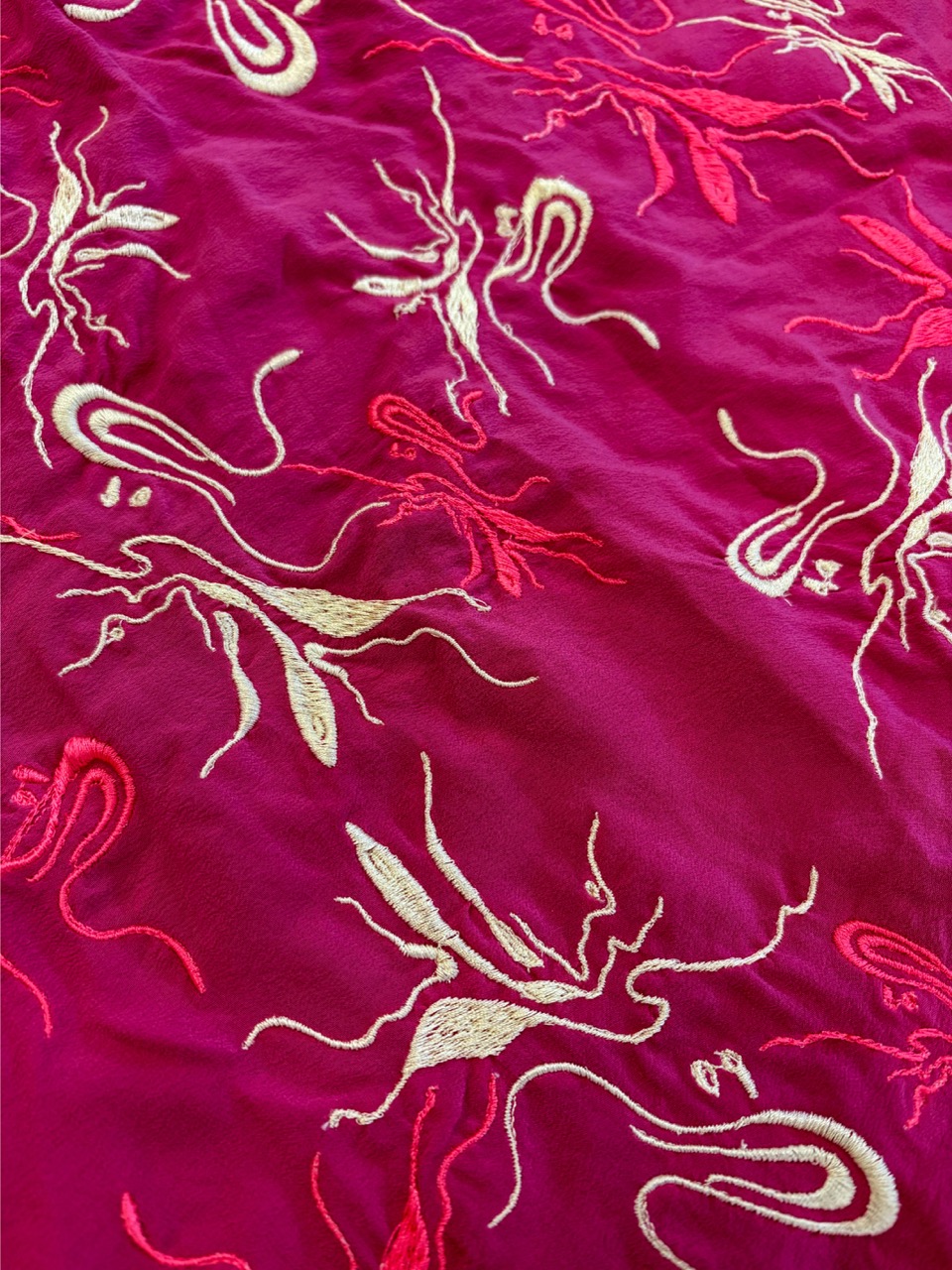 A close-up of a vibrant pink fabric featuring intricate embroidery. The design includes abstract shapes in white and orange, creating a dynamic pattern across the surface.