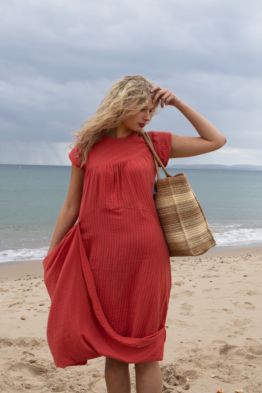 A woman in a coral-coloured dress carrying a straw bag stands on a sandy beach. The ocean is in the background under an overcast sky, with gentle waves visible on the water.