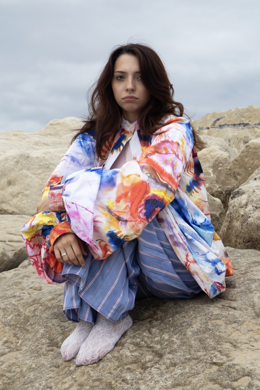 A young woman with long brown hair is sitting on large rocks by the sea, wearing a brightly coloured, patterned jacket and blue striped trousers. She is barefoot, with her arms resting on her knees, against a cloudy sky backdrop.