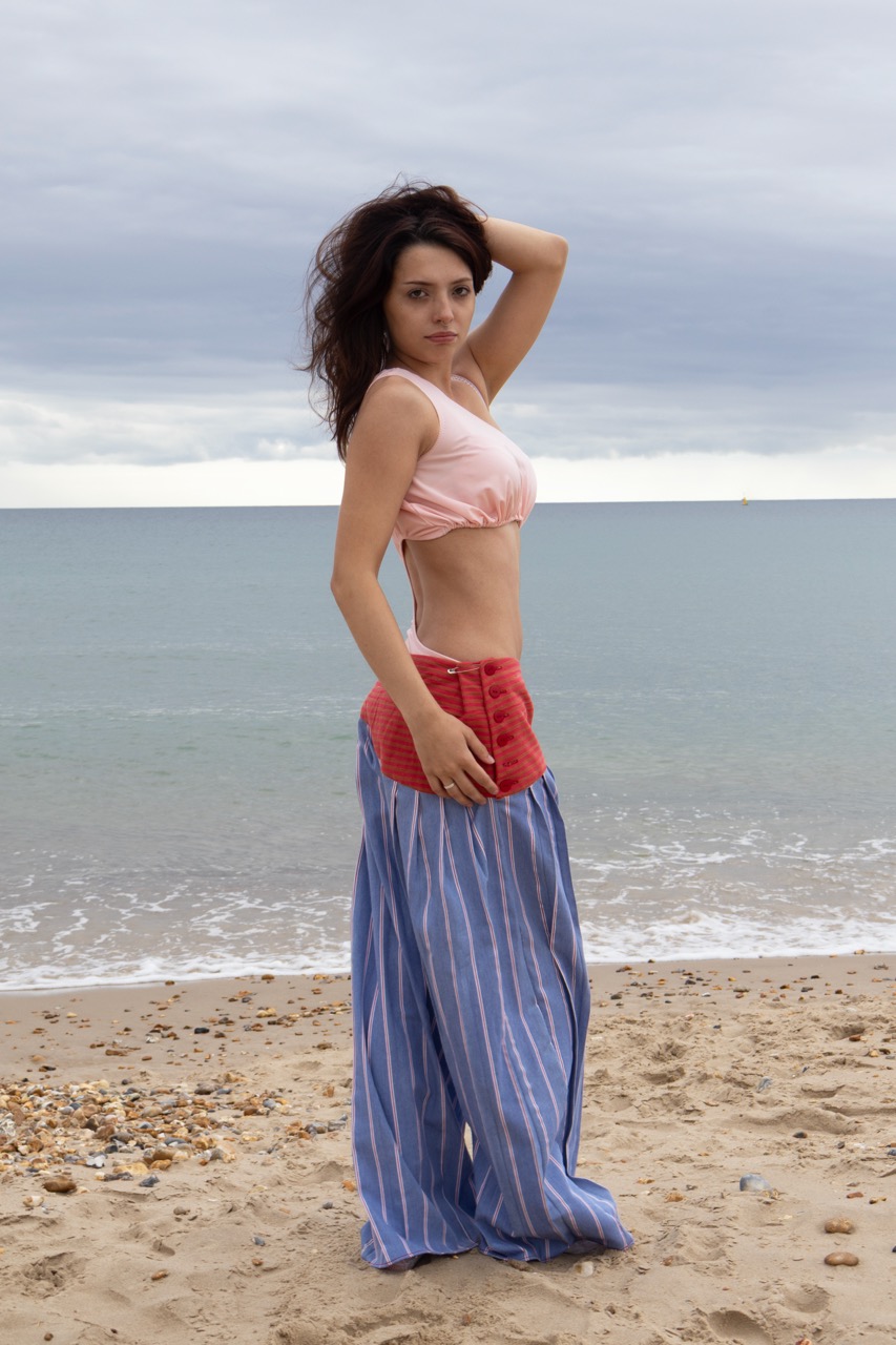 A woman stands on a sandy beach, wearing a pink crop top and loose striped trousers. She poses with one hand on her hip, facing slightly towards the camera, with a calm sea and cloudy sky in the background.