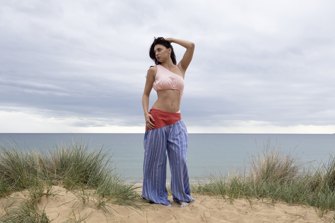 A woman with long dark hair poses on a sandy beach, wearing a pink crop top and loose striped trousers in shades of blue and red. The background features cloudy skies and ocean waves, with tufts of grass surrounding her.