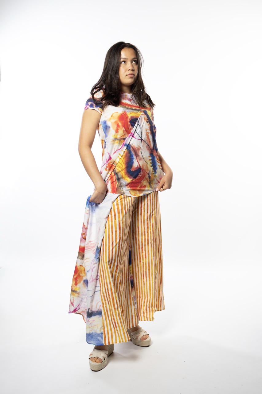 A model stands in a colourful outfit consisting of a patterned top featuring hues of red, yellow, and blue, paired with striped, yellow and white wide-leg trousers. The background is plain white.