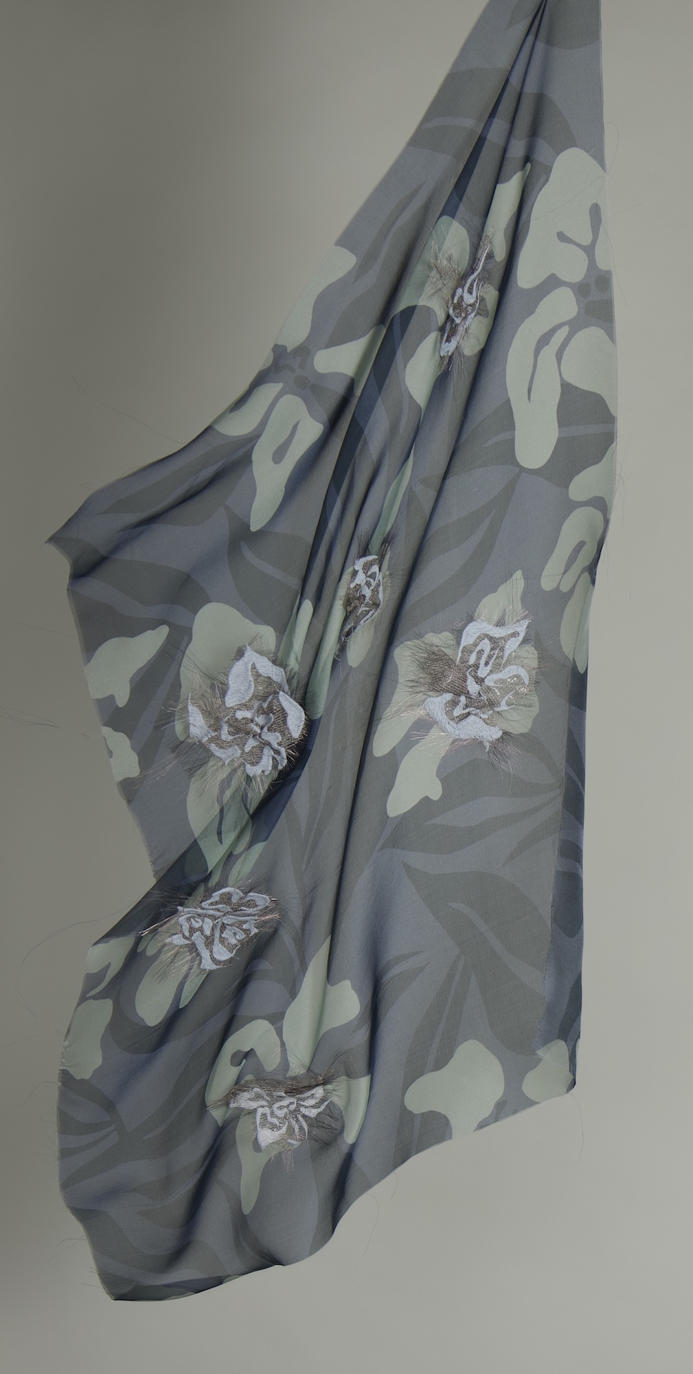 A sheer fabric draped gracefully, featuring a camouflage pattern in muted greens and light greys, with floral designs in whites and browns placed throughout. The background is a plain, light grey wall.