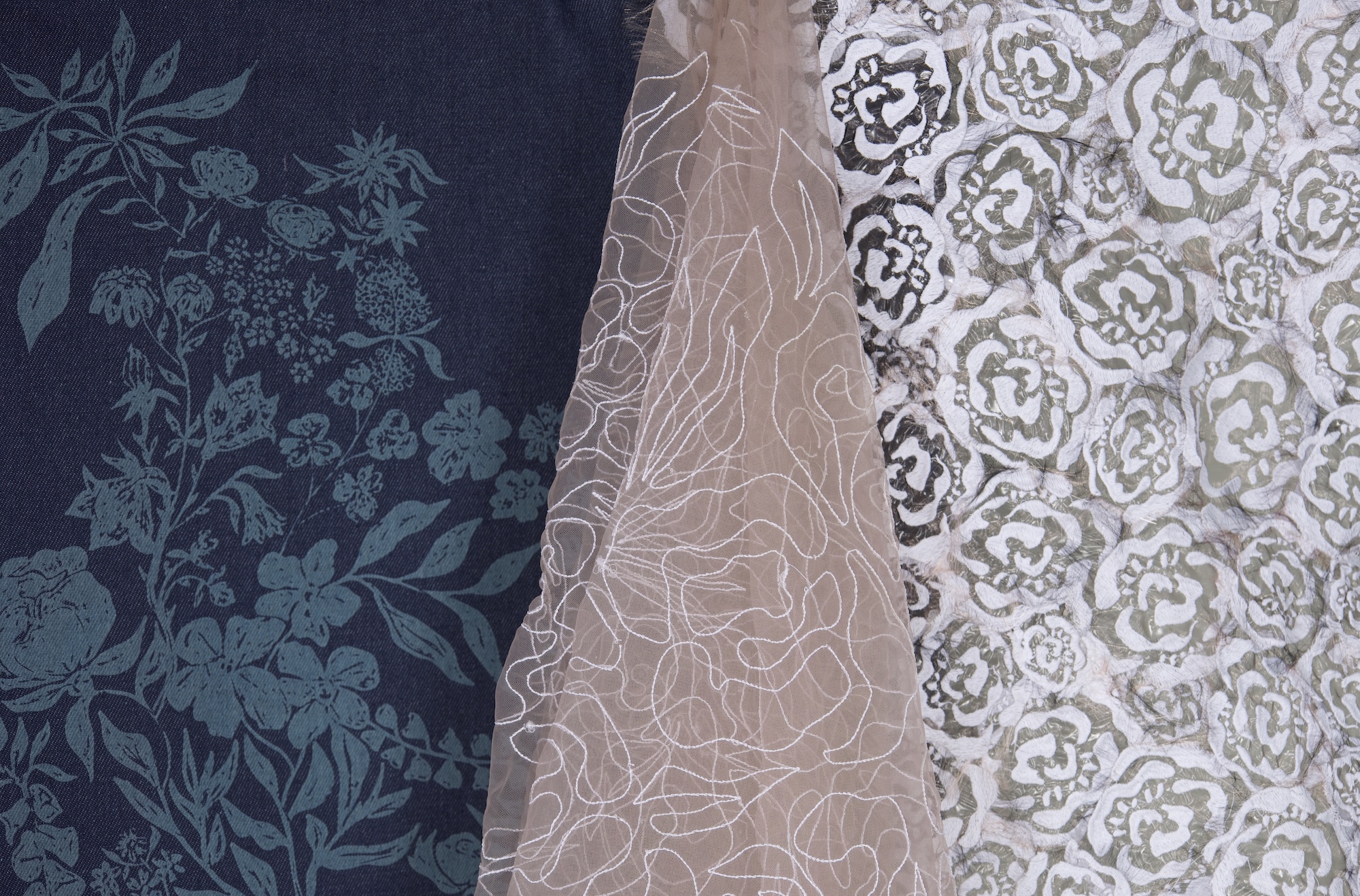 Three distinct fabric samples are displayed side by side. The left sample features a dark navy background with teal floral designs, the centre is a light brown sheer fabric with white floral embroidery, and the right sample has a patterned design of large white flowers on a muted background.