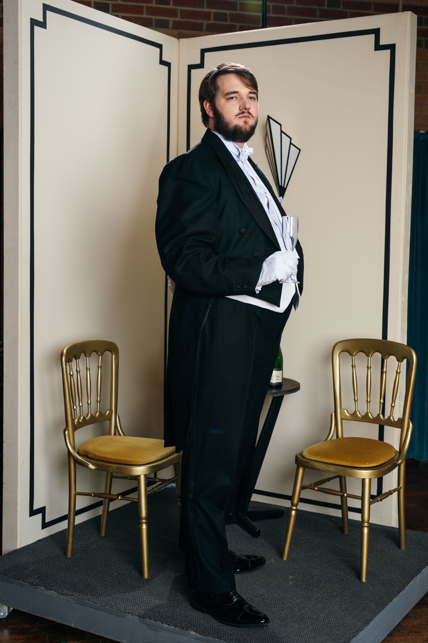 A man in a formal black tuxedo with a bow tie stands confidently beside a small table holding a champagne bottle. Two gold chairs are positioned in front of a decorative backdrop featuring a geometric design.