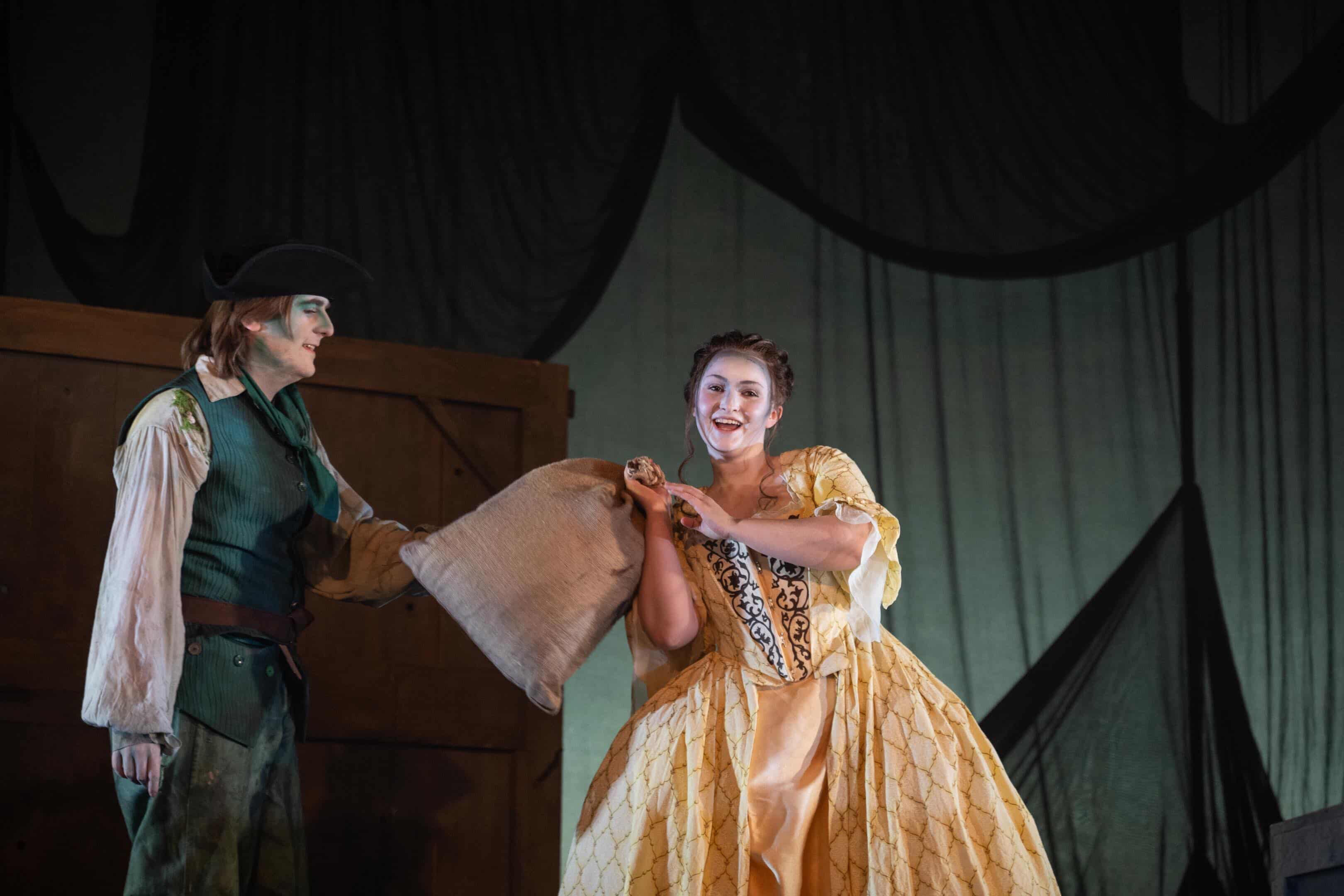 A woman in a yellow gown with decorative detailing holds a large sack while standing next to a man dressed in a green jacket and hat. The backdrop features draped fabric and wooden structures, creating a theatrical setting.
