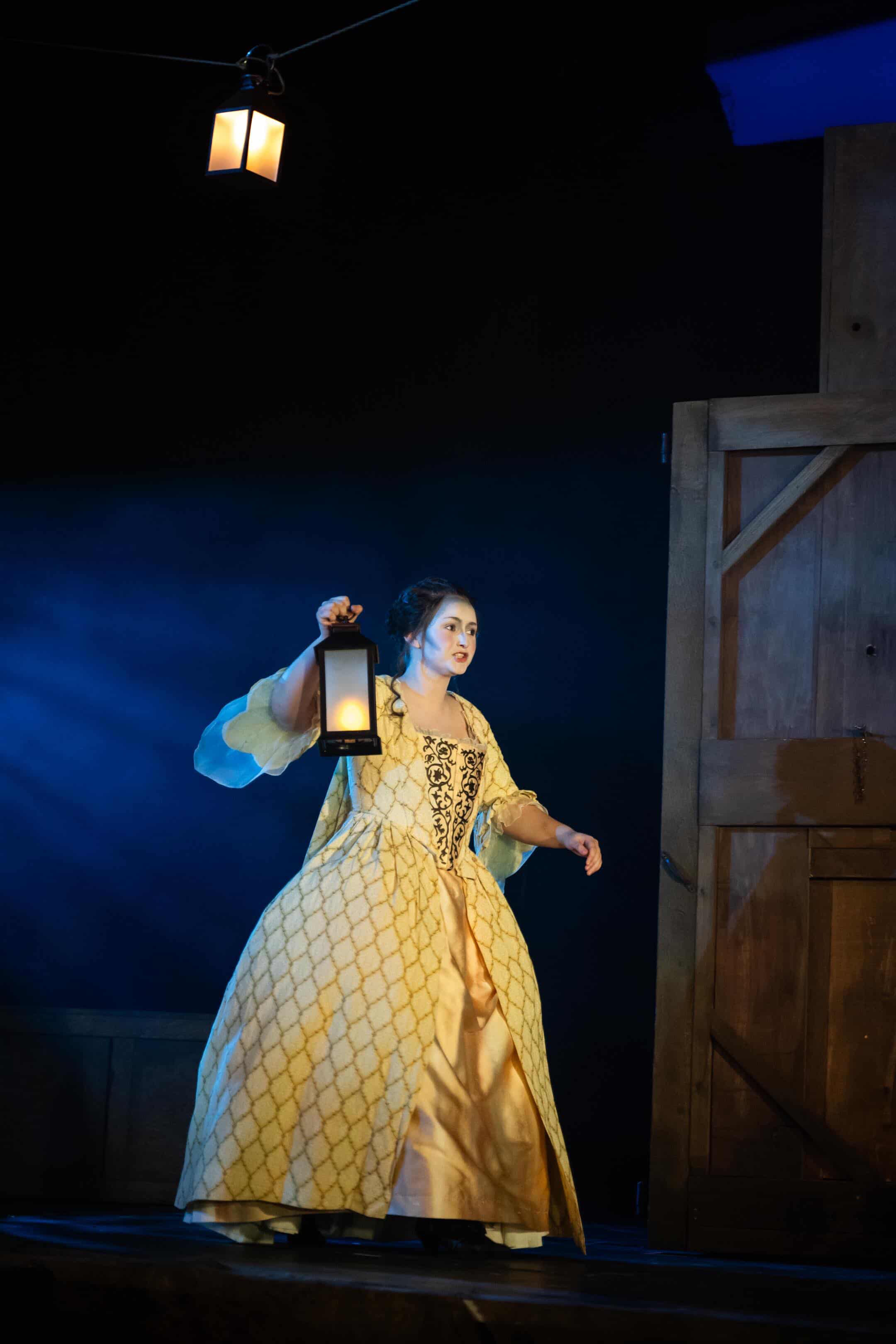 A woman dressed in a yellow gown with a diamond pattern holds a lantern in her left hand. She stands on stage, with a wooden doorway to her right and a lantern hanging from above, while blue lighting creates a dramatic effect.