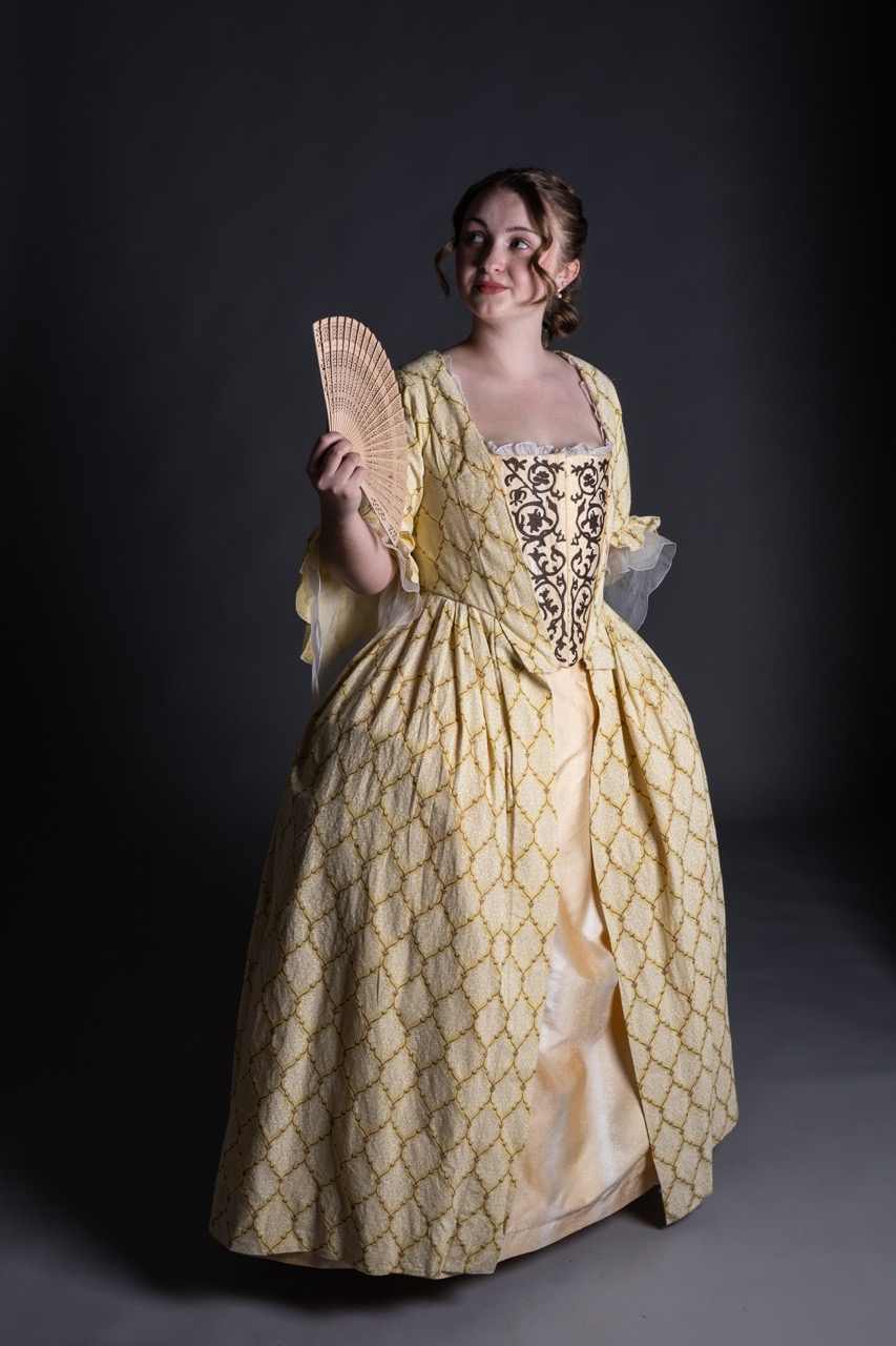 A woman wearing a yellow gown with intricate patterns and puffed sleeves holds a fan in her right hand. She stands against a dark background, with her hair styled in loose curls, and a lace trim visible at the neckline.