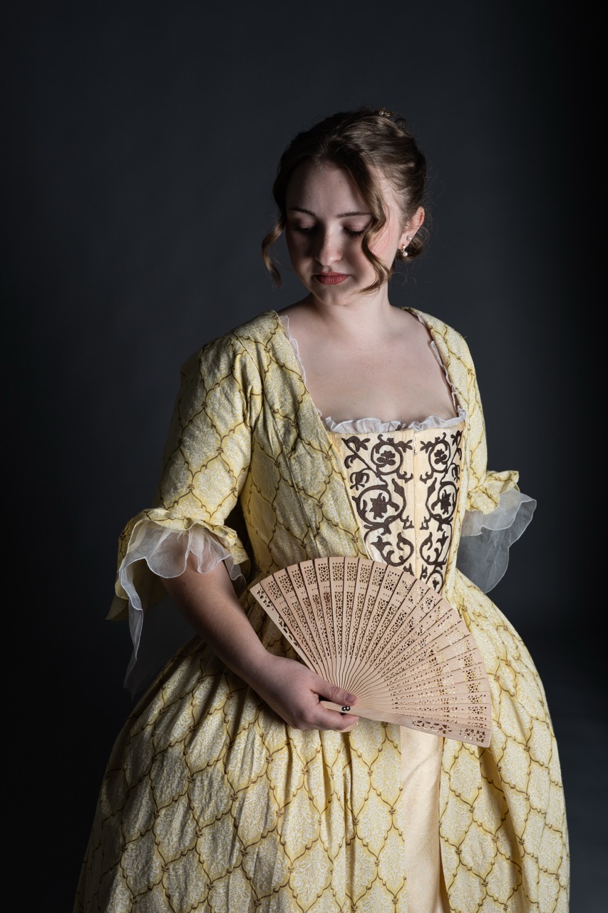 A woman dressed in an elaborate yellow gown with intricate patterns stands gracefully, holding a decorative fan. The gown features light ruffled sleeves, and the background is a shaded grey, enhancing the focus on her elegant attire.