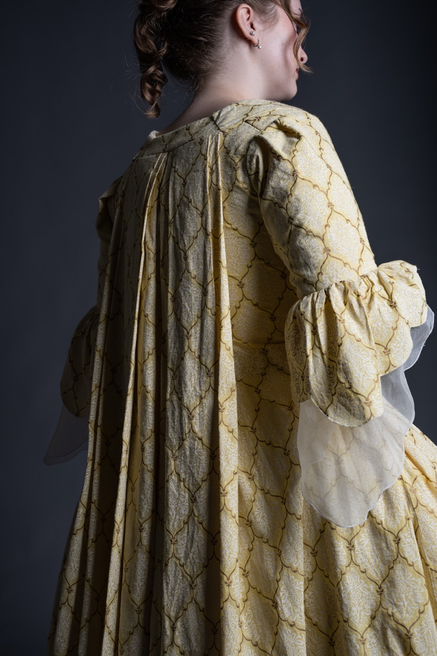 A woman wearing a yellow dress with a geometric pattern and puffed sleeves stands in profile. The dress features a row of pleats down the back, and the setting has a dark background that contrasts with the lighter fabric.