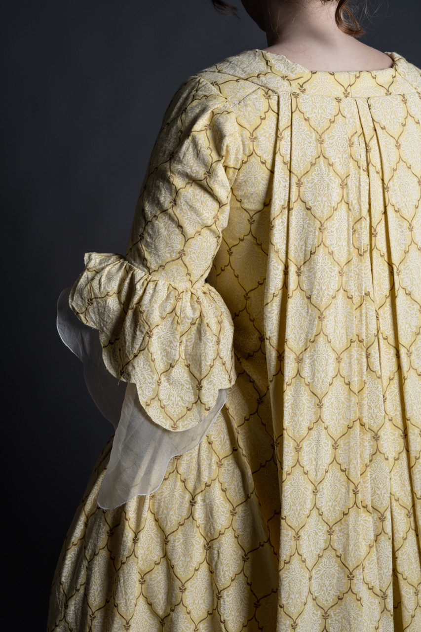 A woman with light brown hair is wearing a vintage yellow dress with a textured pattern, featuring puffed sleeves and a high neckline. The image focuses on the back view of the dress against a dark background.