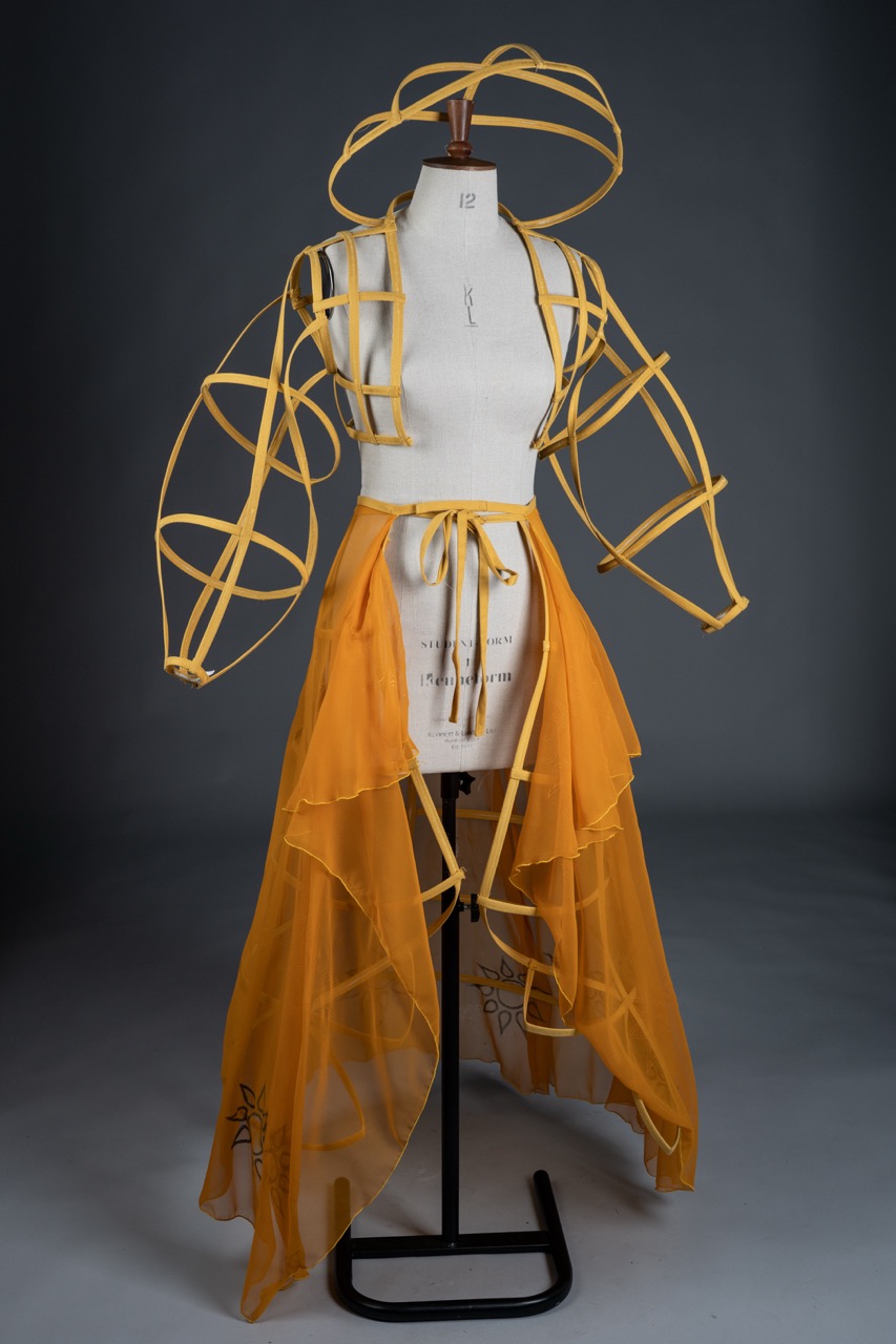 A fashion design displayed on a mannequin, featuring a yellow garment with a sheer, layered skirt and structured, open sleeves. The outfit consists of yellow ribbons forming a geometric design, contrasting with the flowing fabric of the skirt.