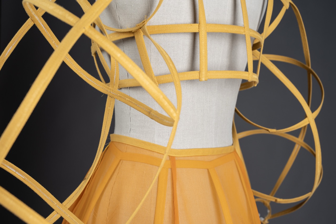 A close-up of a fashion design on a mannequin featuring a structured yellow top made of interwoven straps. The lower part is a yellow skirt with a pleated design, set against a dark background.