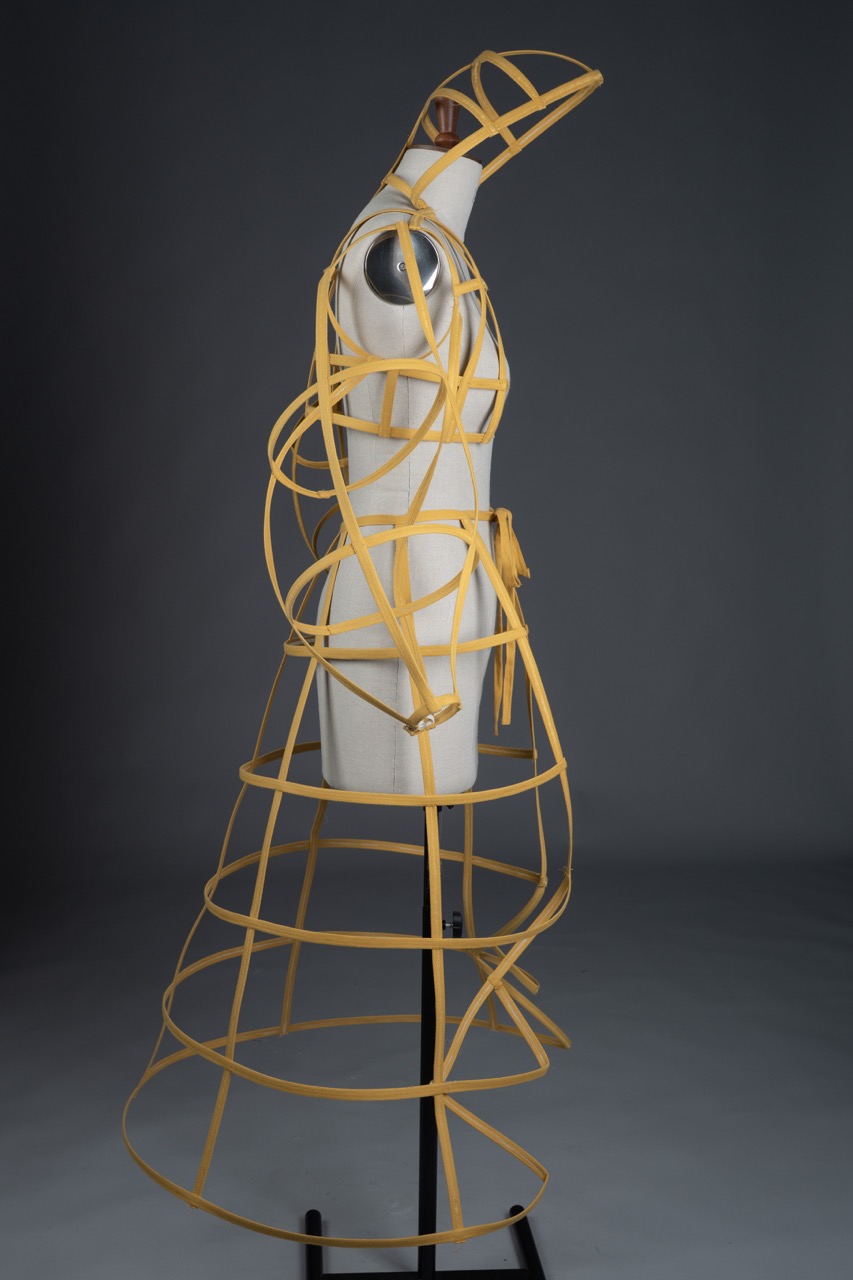 A dressmaking mannequin covered in yellow structured ribbon-like material, creating a geometric and open design. The mannequin is positioned on a black stand against a grey background, showcasing the unique fashion display.