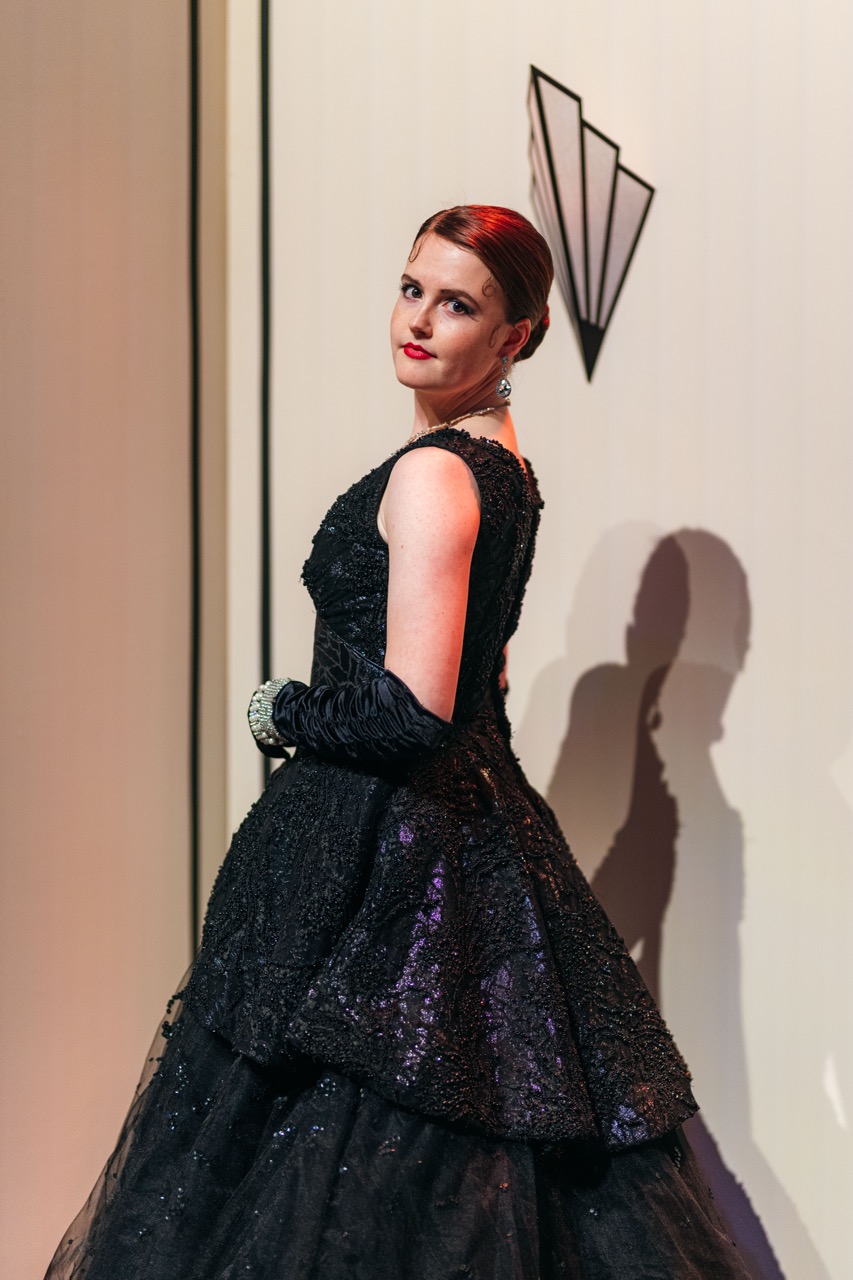A woman wearing a black gown with a full skirt and matching gloves stands in profile against a pale background. The gown features intricate detailing, and she has her hair styled elegantly. A geometric design is visible on the wall behind her.