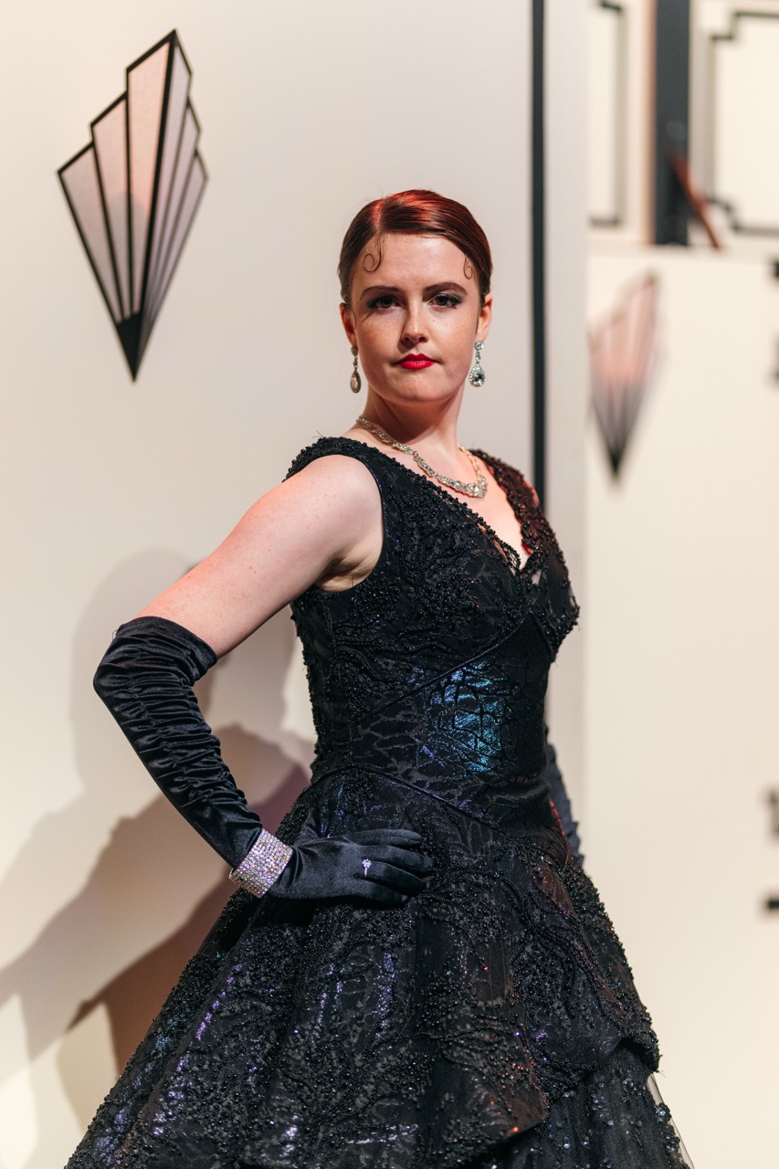 A woman with red hair styled in vintage waves wears a black evening gown with intricate detailing and long black gloves. She stands in front of a patterned backdrop featuring art deco motifs, with a confident pose.