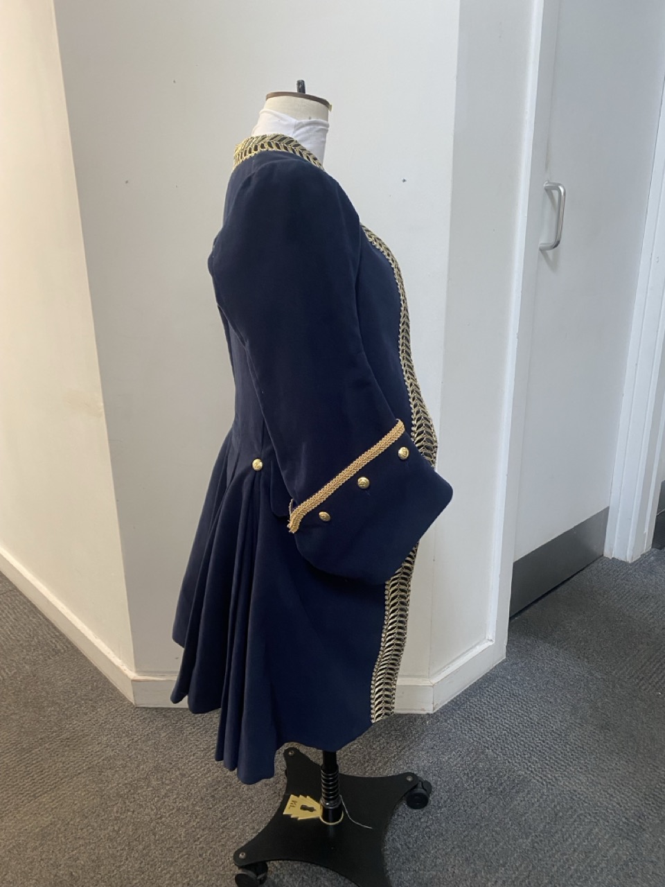 A navy blue coat displayed on a mannequin, featuring gold buttons and decorative trim along the edges. The coat has a fitted silhouette with a flared skirt section and is set against a simple interior background with white walls and grey carpeting.