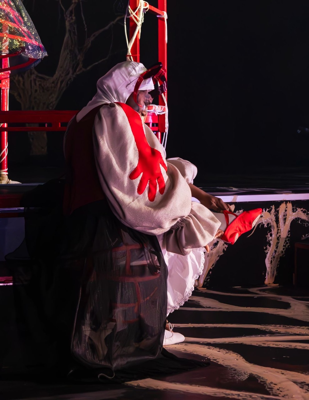 A performer dressed in a whimsical costume, featuring a white hood and red oversized hands, sits on a stage, adjusting bright red shoes. The background includes abstract, dark scenery with illuminated details.