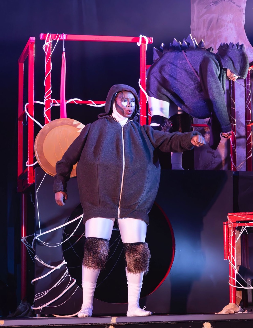 A performer in a large grey outfit with a hood and furry details stands on stage, pointing. Behind them, another performer is dressed in dark attire, manipulating a structure made of red and black materials with lighting effects in the background.