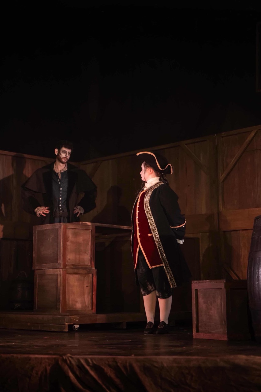 Two male actors are performing on stage in a theatrical setting. One actor, wearing a black cloak, stands beside a wooden table, while the other, dressed in a red coat and tricorn hat, faces him. The backdrop features wooden panels, contributing to a dramatic atmosphere.