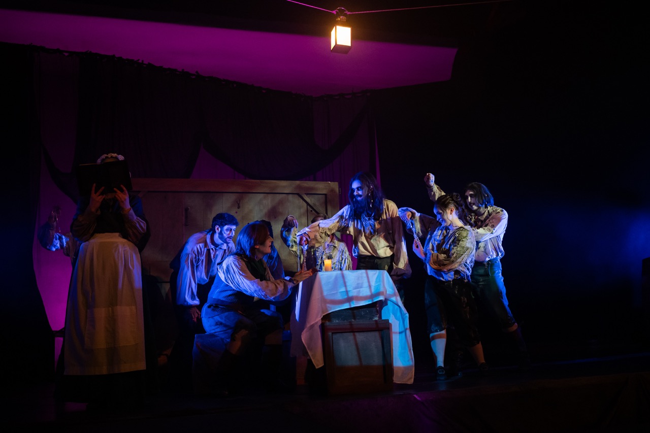 A theatrical scene featuring seven actors in period costumes, gathered around a table set with bottles and a glass. The stage is dimly lit with purple and blue lighting, creating contrast with the wooden backdrop and shadowy corners.