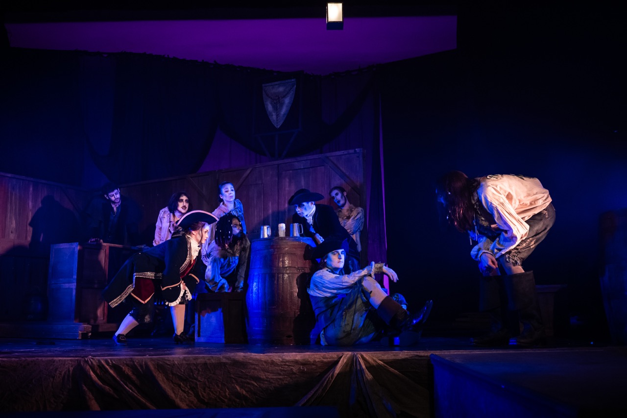A theatrical stage scene featuring actors in period costumes, including a pirate-themed outfit. Some actors are seated on barrels, while others stand or lean, surrounded by wooden sets and dim theatrical lighting.