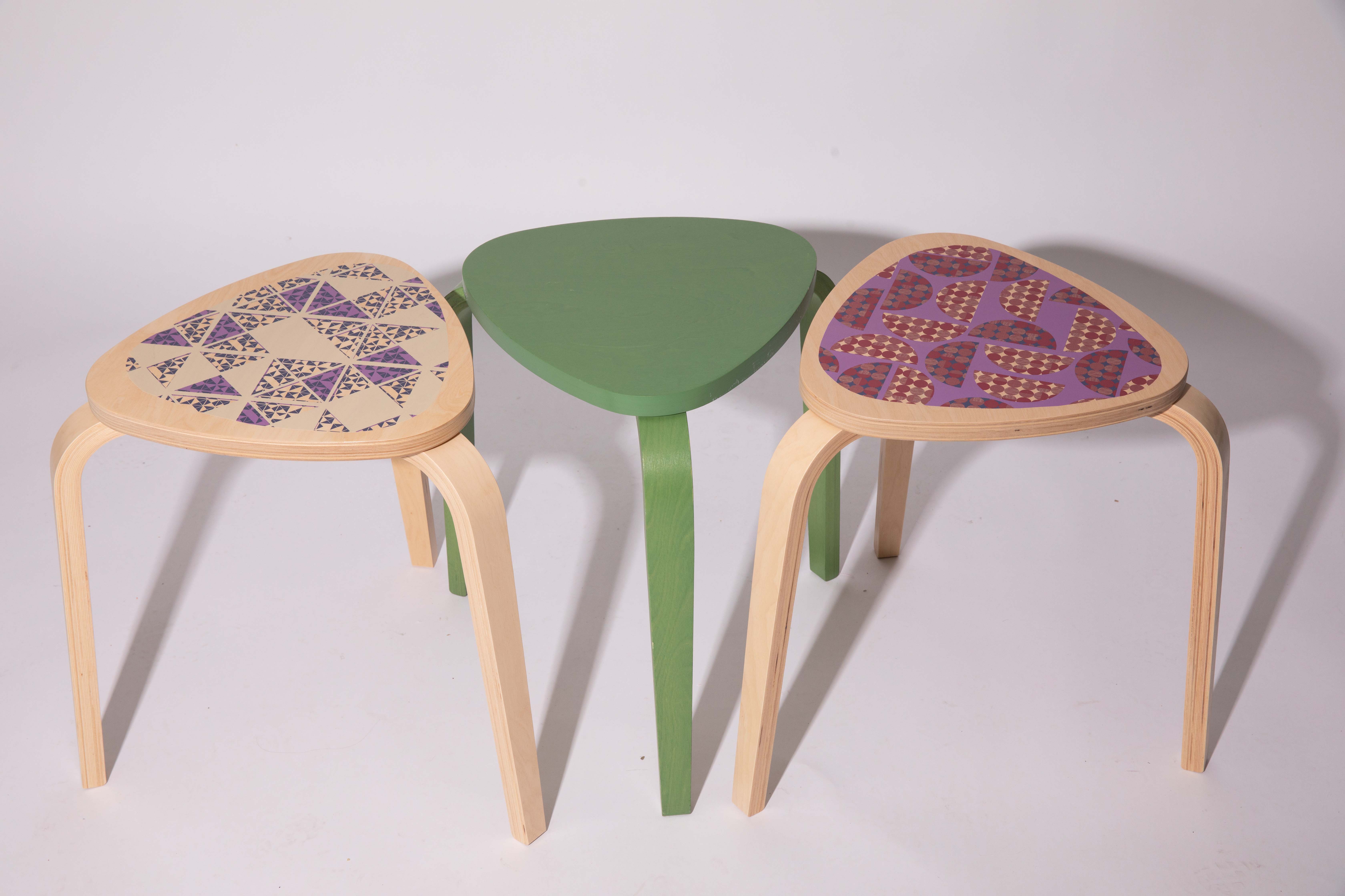 Three triangular stools arranged together on a light background. The left stool has a patterned seat in purple and beige, the center stool is painted green, and the right stool features a purple and orange geometric design.