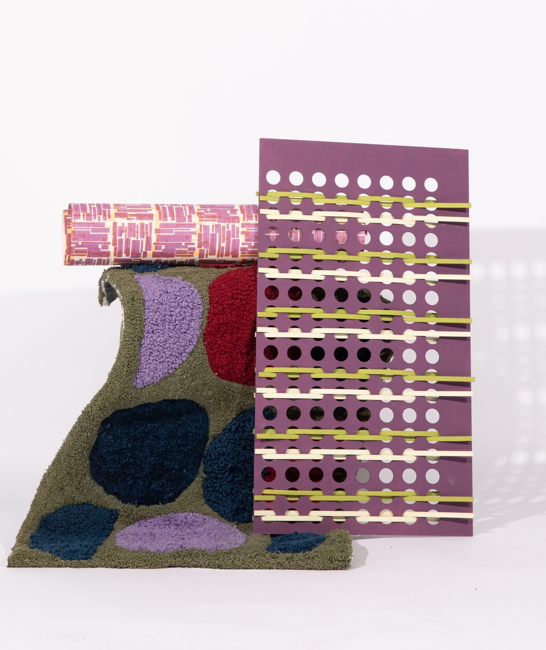 A textured piece of fabric in green, featuring abstract shapes in purple and red, stands upright. Behind it is a purple panel with white and black circular cut-outs, covered by a pink patterned fabric roll with geometric designs.