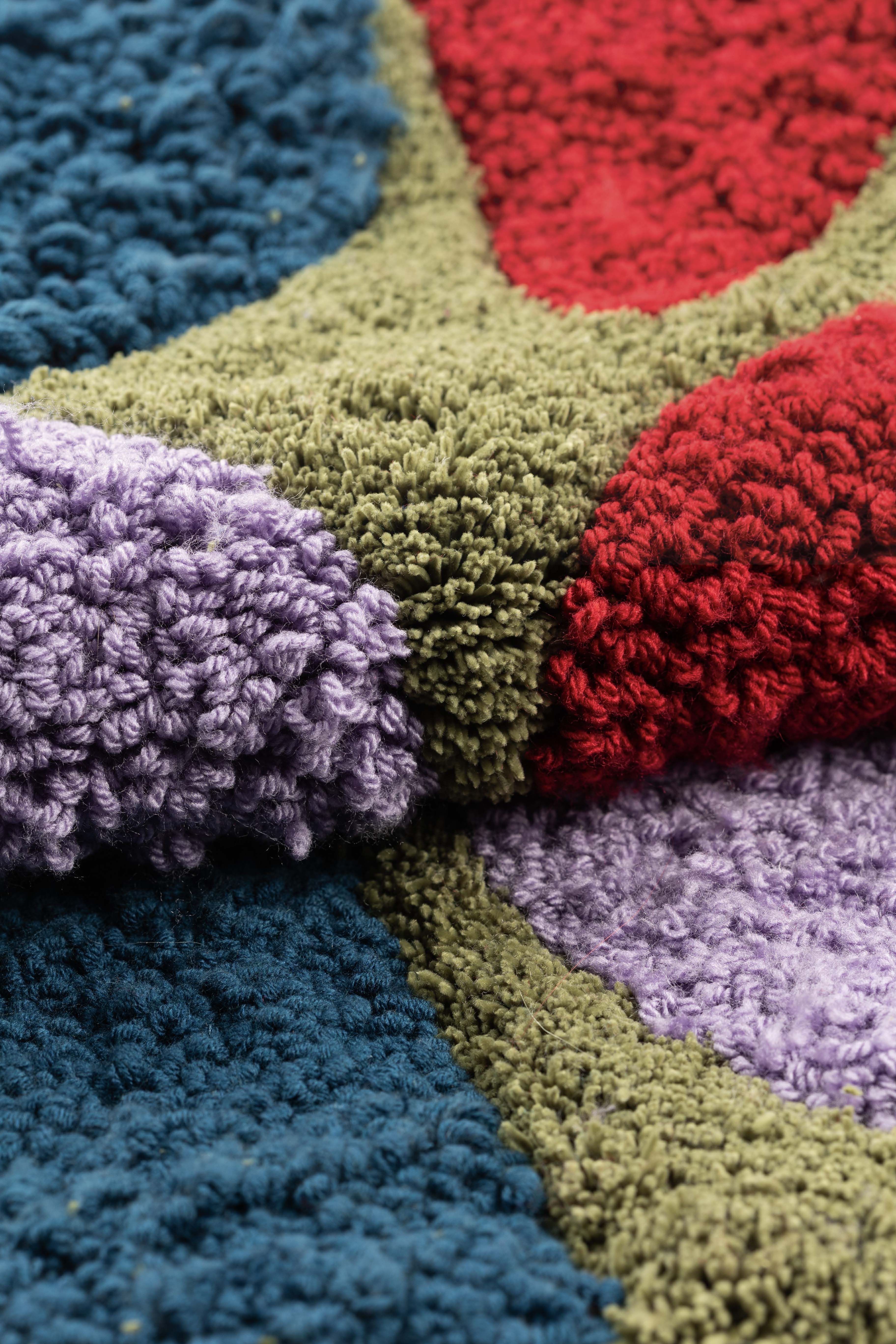 A close-up of a textured rug featuring a pattern with segments in blue, green, red, and purple. The rug displays a shaggy, plush texture, with each coloured area seamlessly blending into the next.
