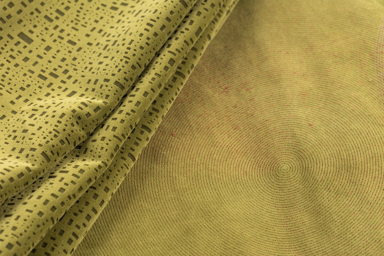 A close-up of two pieces of fabric in varying shades of green. One piece features a patterned design with small black squares, while the other has a circular texture, both laid on a surface that showcases their contrasting designs.