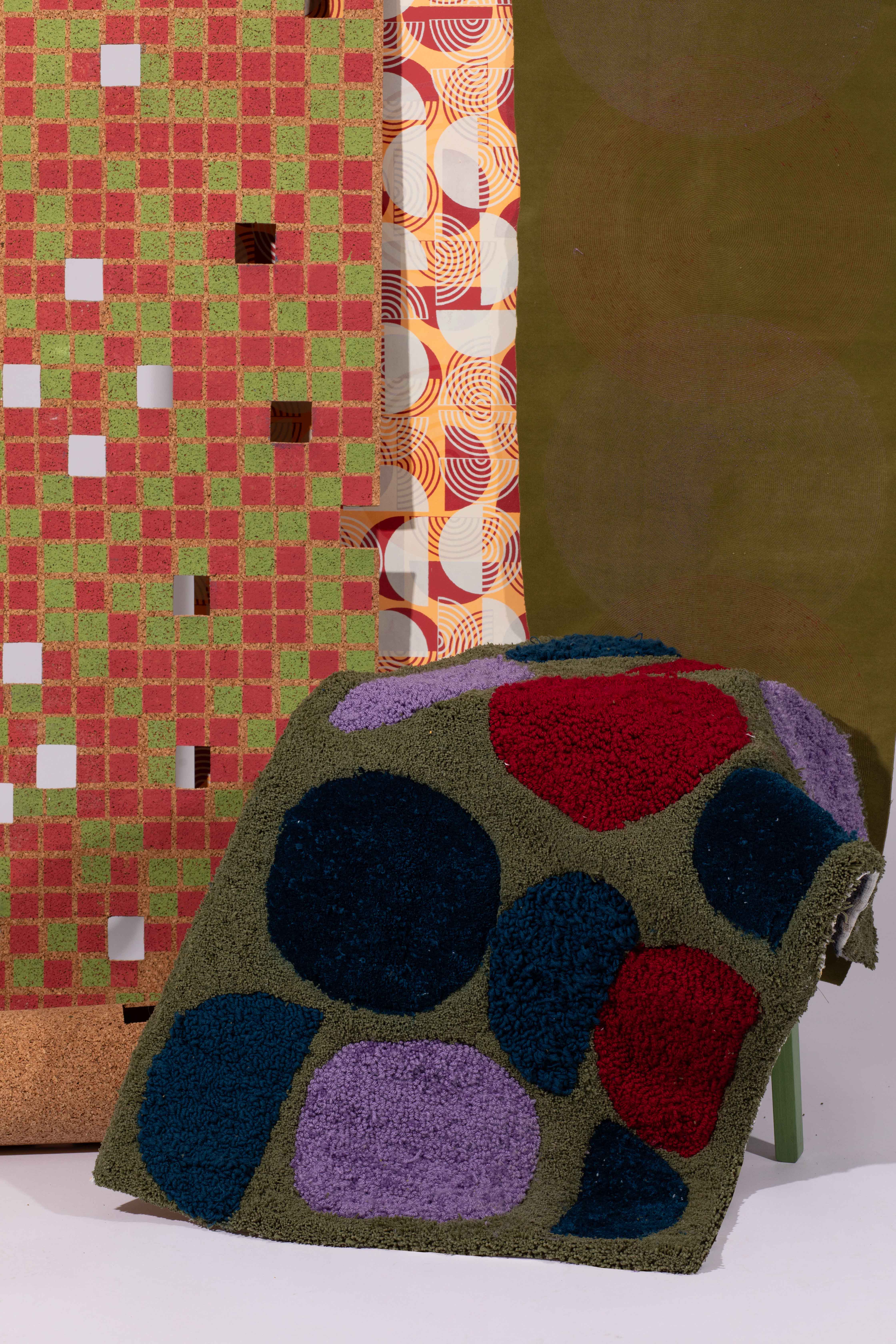 An assortment of patterned textiles. In the foreground, a green textured fabric with large blue, purple, red, and black circles drapes over a surface. Behind, two patterned fabrics feature a grid design in red and green, and a swirling design in orange, cream, and brown.
