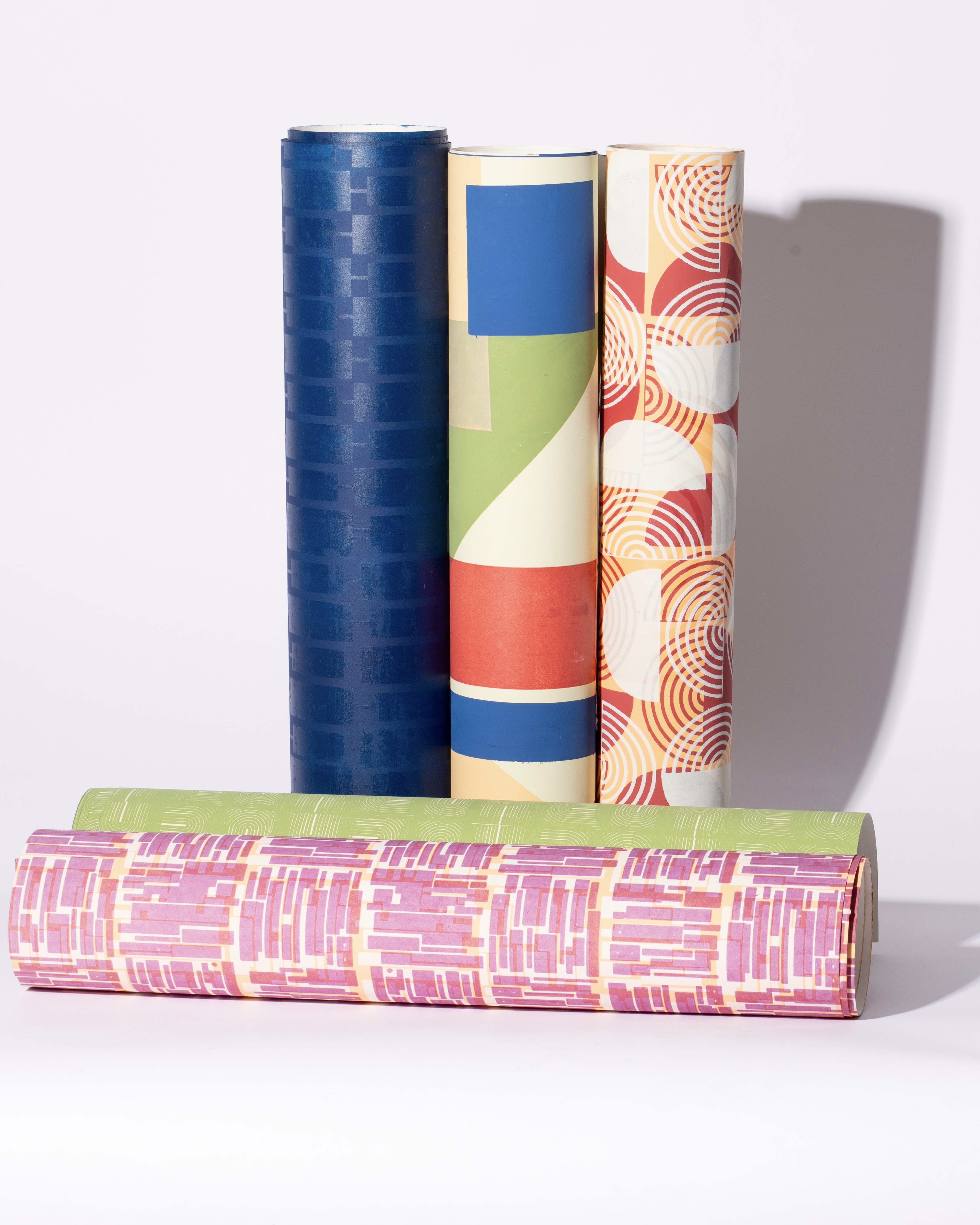 Five rolls of decorative wrapping paper stacked vertically, featuring various colourful patterns. The top two rolls are in shades of blue and patterned with circles and lines, while the bottom three rolls include pink, green, and abstract designs.