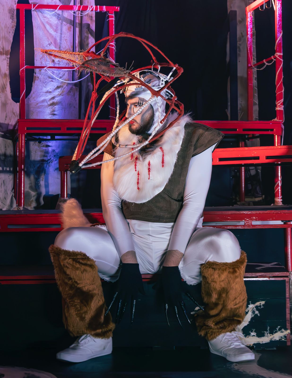 A performer dressed in a unique costume resembling a deer, with antlers and fur details. They are seated with a dramatic pose on a stage, surrounded by red structures and dim lighting.