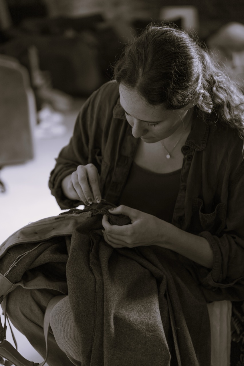A young woman in a dark shirt is focused on sewing fabric while seated. Her hair is styled in loose waves, and she is using her hands to work carefully on a piece of clothing in a softly lit room with soft background details.