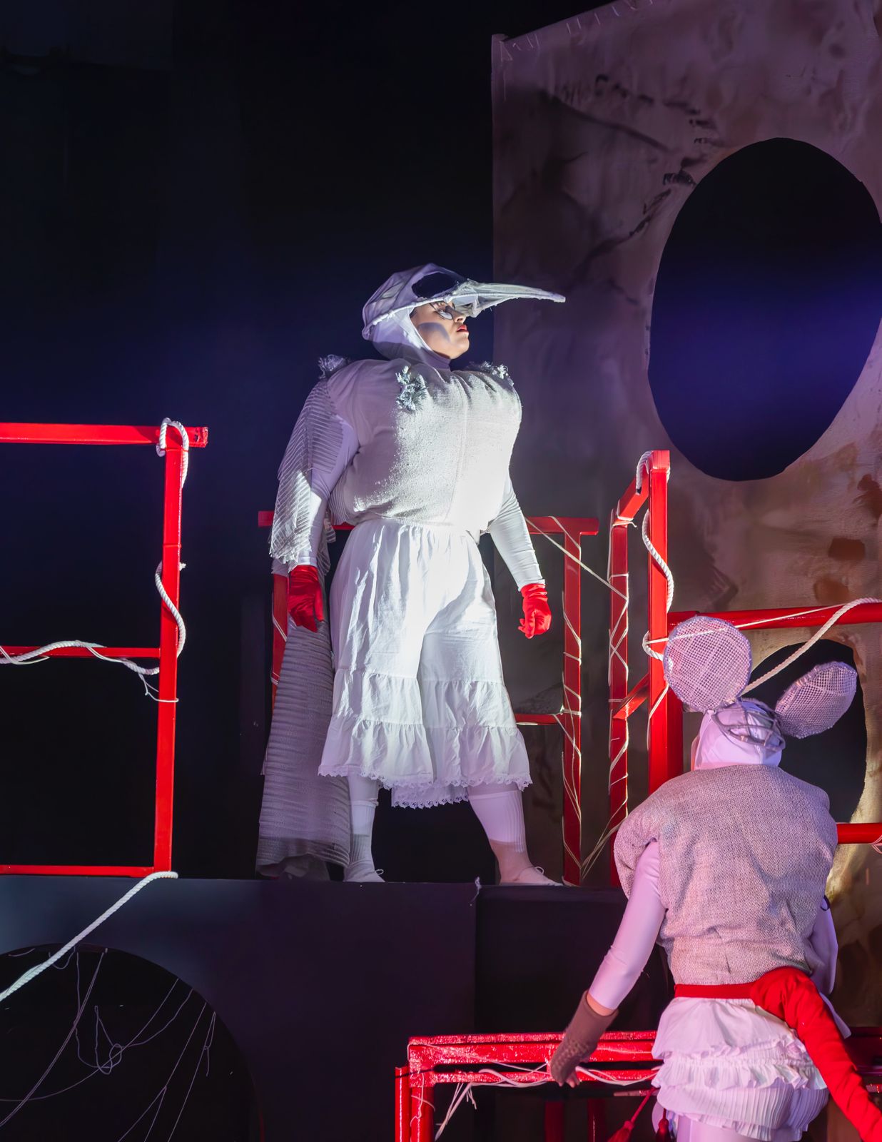 A performer dressed in a white outfit with a beak-like mask stands on a stage with red railings, while another performer in a mouse costume, wearing a tail and ears, is partially visible in the foreground. The background includes abstract shapes and dramatic lighting.