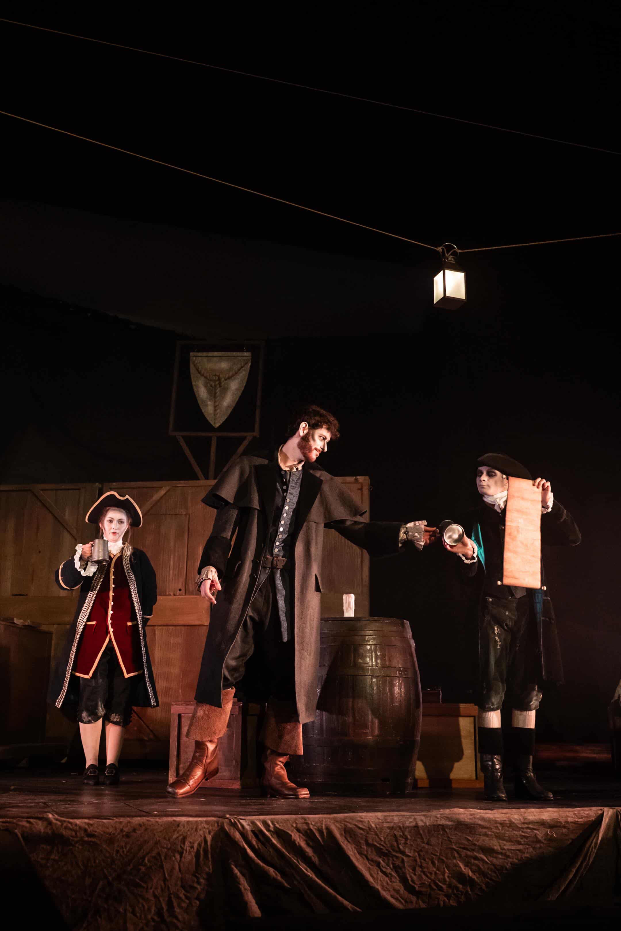 Three actors are performing on stage. One actor, dressed in a long black coat and holding a drink, faces another in a black hat holding a scroll. A third actor, in a red coat and white face paint, stands nearby. The stage is set with barrels and wooden props.
