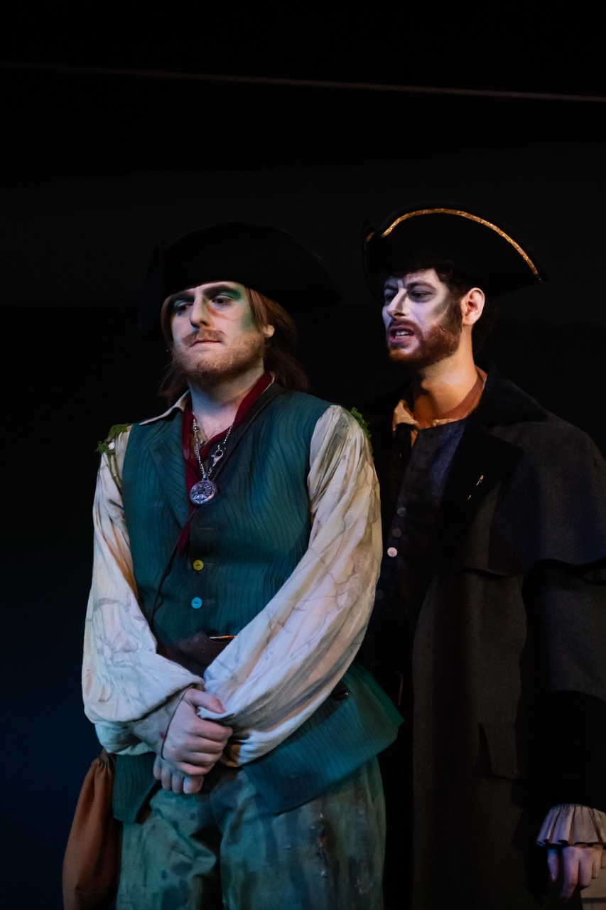 Two actors are on stage in period costumes. The first actor, dressed in a blue vest over a white shirt, has long hair and a black hat. The second actor wears a dark coat and hat, with dramatic makeup on his face. A dark backdrop is visible behind them.
