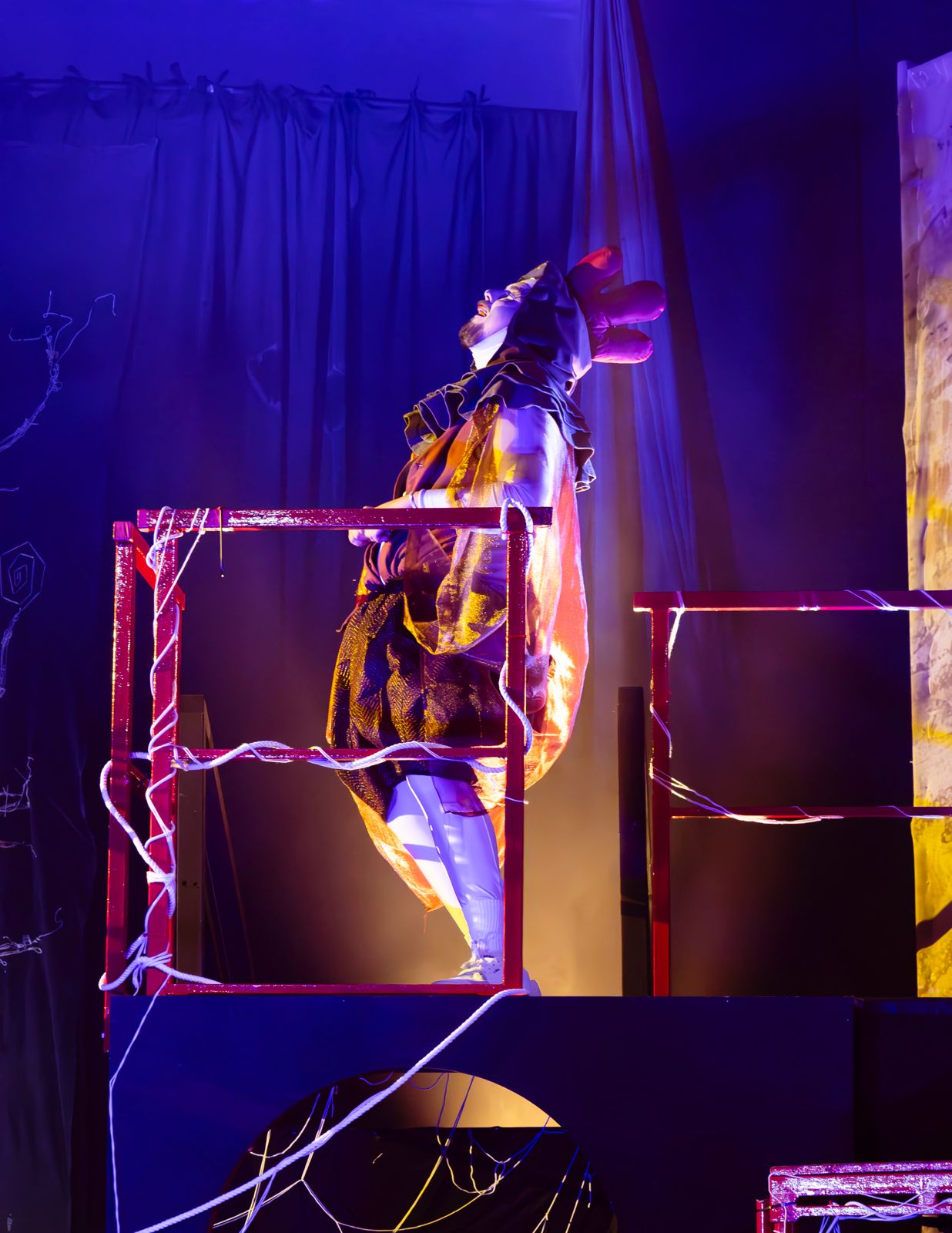 A performer wearing a costume with feathers stands on a red platform in a dramatic pose, surrounded by purple and blue lighting. The backdrop features dark curtains and abstract designs.