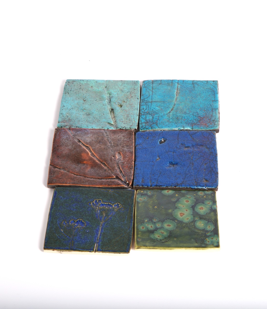 Six square ceramic tiles arranged in a two-by-three grid, featuring various shades of blue and green. Each tile has a distinct texture and finish, with some displaying patterns and markings.