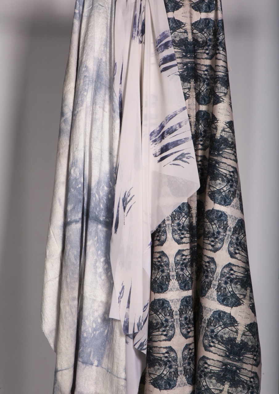 Three pieces of fabric hanging vertically, displaying different patterns. The left fabric features a pale blue abstract design, the centre fabric is a plain light shade, and the right fabric showcases a dark circular pattern on a light background.