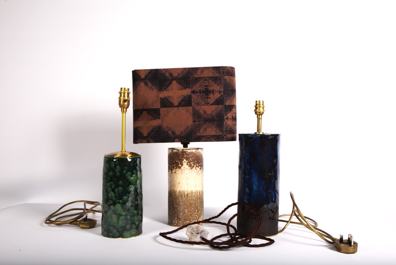 Three table lamps on a white surface. The left lamp has a textured green base, the middle lamp features a beige base with a patterned brown shade, and the right lamp has a dark blue base, both with bronze fittings and cords.