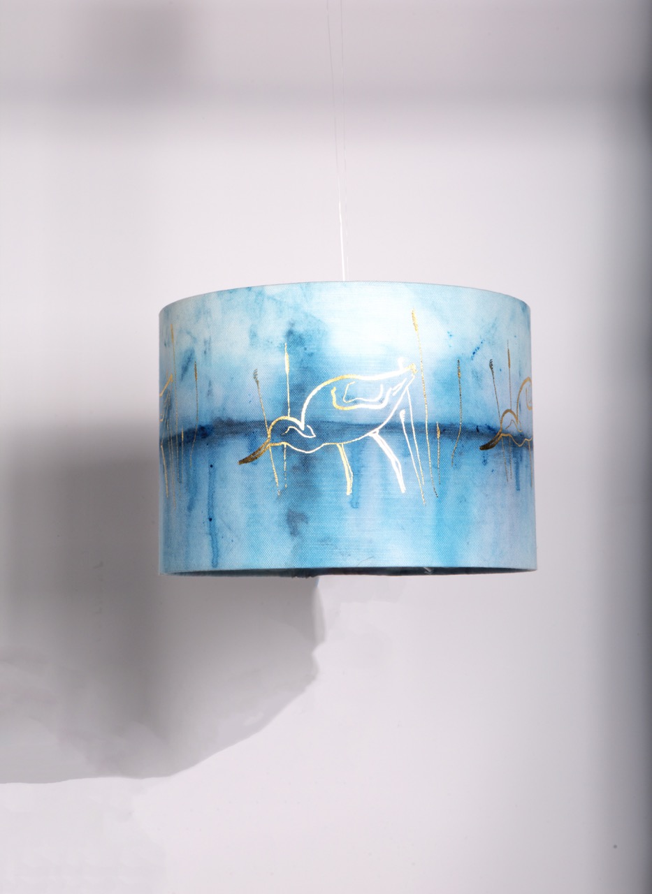 A round lampshade with a blue and white watercolour pattern featuring a graceful bird design in gold. The lampshade is suspended from the ceiling, with a plain white background enhancing its artistic appearance.