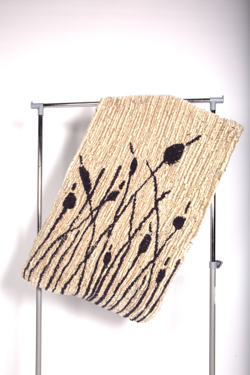 A woven beige mat with a black floral design displaying stems and abstract leaves. The mat is hanging on a silver rack.