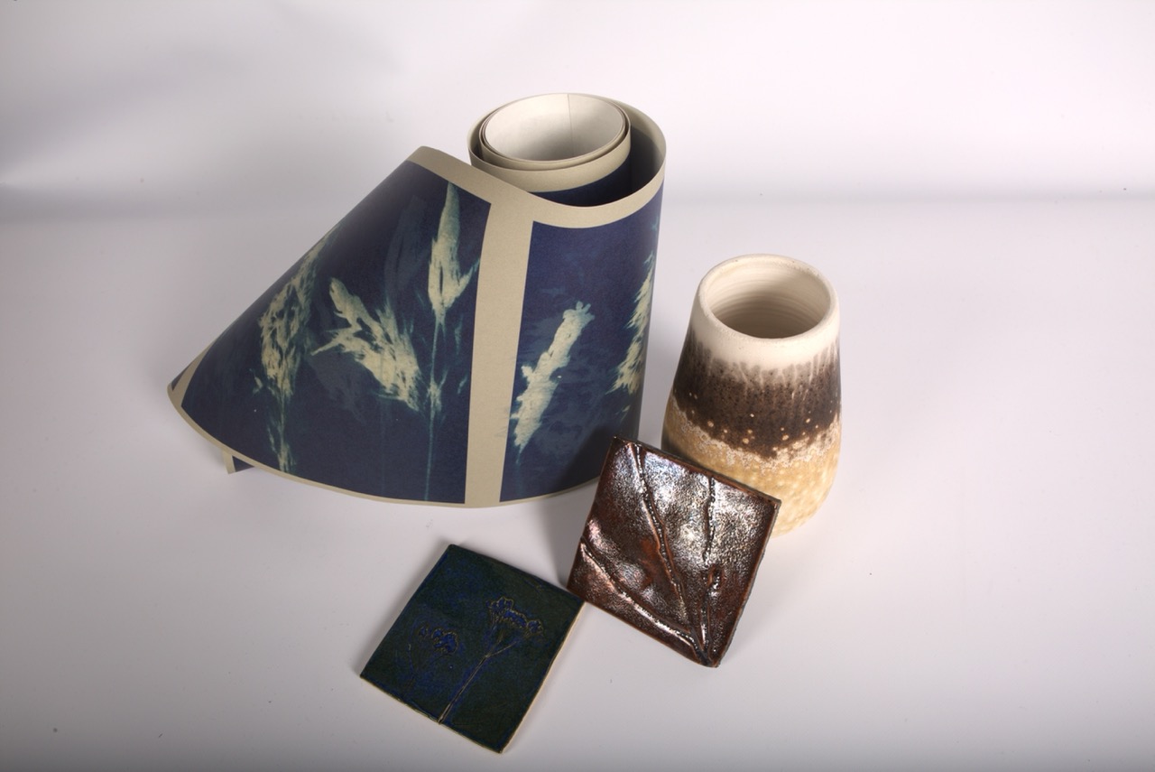 A rolled piece of blue paper featuring plant designs rests beside two ceramic vases: one with a textured surface and gradient brown to cream colour, the other a plain white cylinder. Also, there are two square ceramic tiles with glossy finishes.