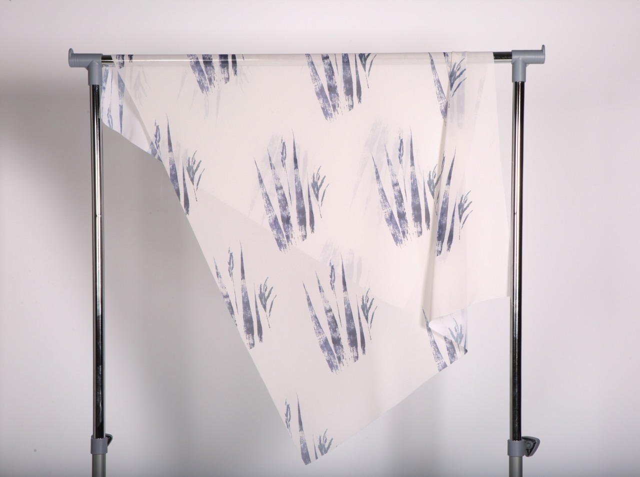 A piece of fabric with a white background featuring a pattern of abstract blue and grey plant silhouettes, draped over a metal rack. The fabric is arranged in a way that creates folds and angles.