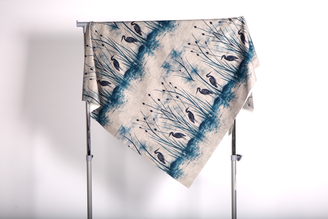 A patterned fabric draped over a metal frame. The textile features a design of blue-toned grasses and slender birds on a light background, creating a decorative appearance.