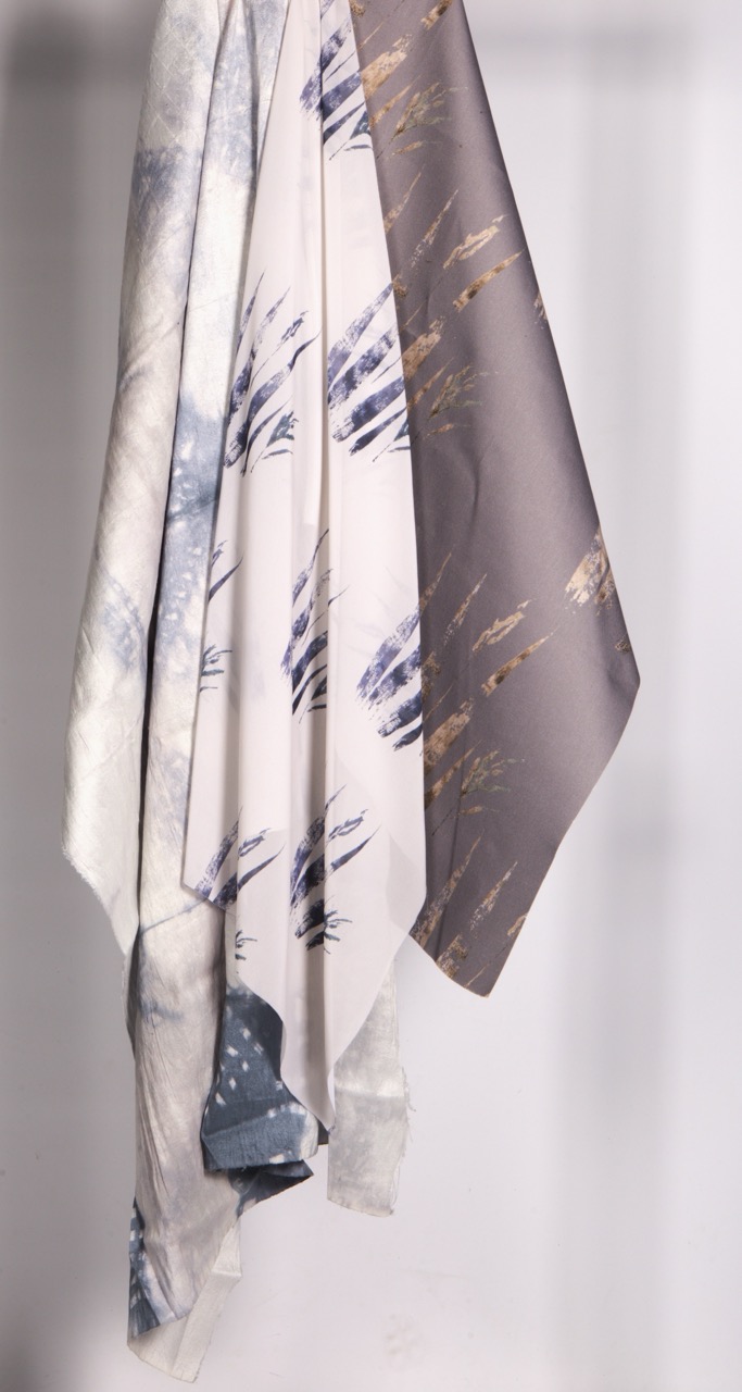 Three pieces of fabric hanging vertically, featuring different patterns. The left fabric has a light pattern with blue and grey hues, the centre fabric is beige with metallic accents, and the right fabric is solid grey. The background is a plain light colour.